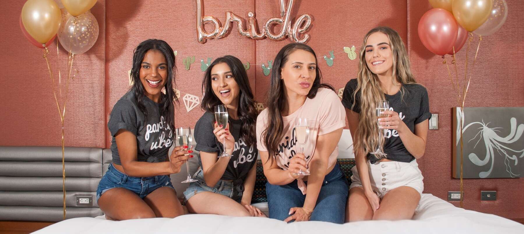 Bachelorette party with a group of four women lounging on a bed under bride decor on the wall.