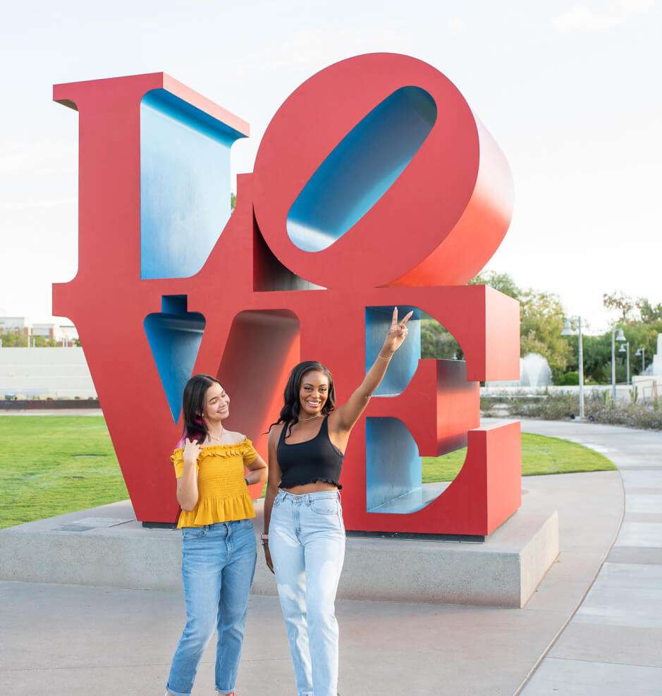 Two women pose by a large love sculpture.