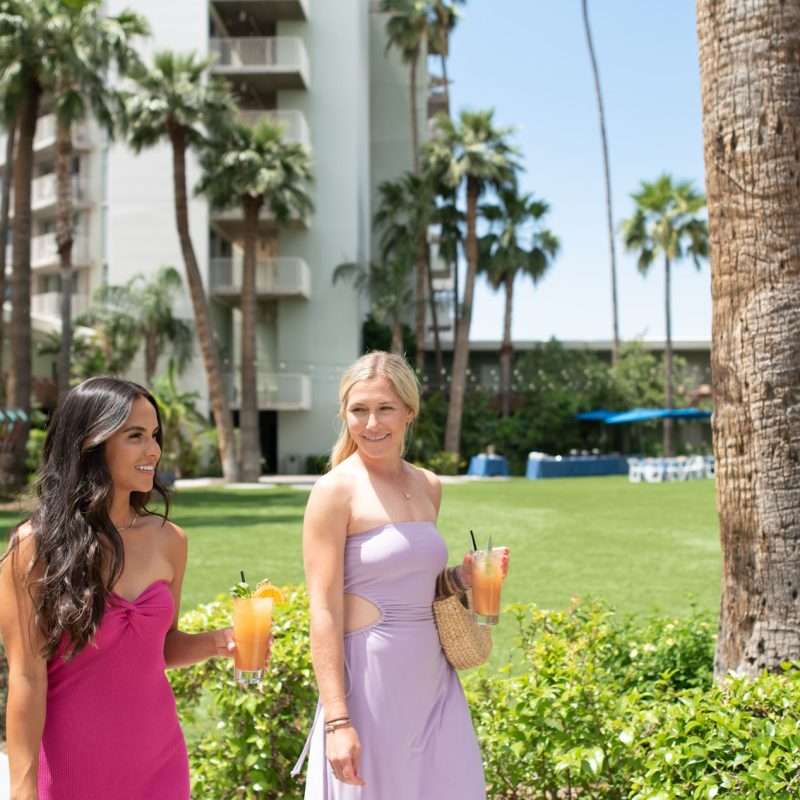 Two women in dresses stroll along a palm-lined path at Hotel Valley Ho.