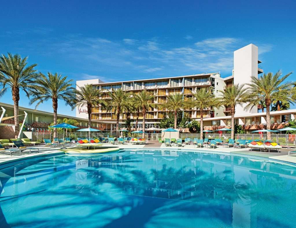 Circular pool outside surrounded by chairs, umbrellas, and trees with a building in the back.